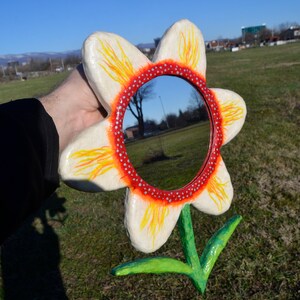 May include: A whimsical, handmade flower mirror. The mirror is round, surrounded by a red and white dotted border, and set within a large, white flower with yellow and orange accents. The stem and leaves are green.