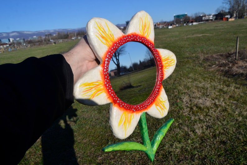May include: A whimsical, handmade flower mirror. The mirror is round, surrounded by a red and white dotted border, and set within a large, white flower with yellow and orange accents. The stem and leaves are green.