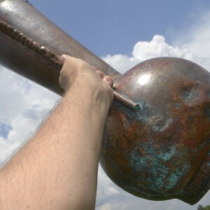 May include: A large, copper vase with a green patina. The vase is being held up by a person's arm.