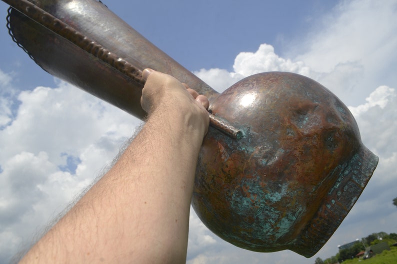 May include: A large, copper vase with a green patina. The vase is being held up by a person's arm.
