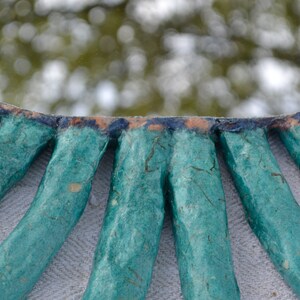 May include: A handmade mirror with a unique, organic design. The mirror's frame is crafted from textured, teal-colored material, radiating outwards from the mirrored surface. The edges are accented with dark blue and orange hues, creating a striking contrast.