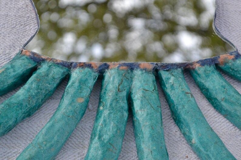 May include: A handmade mirror with a unique, organic design. The mirror's frame is crafted from textured, teal-colored material, radiating outwards from the mirrored surface. The edges are accented with dark blue and orange hues, creating a striking contrast.
