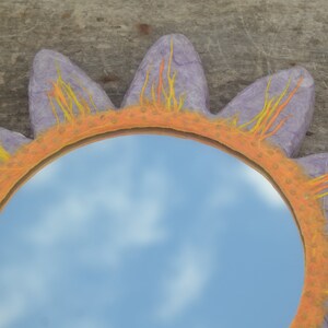 May include: A decorative sun-shaped mirror with a lavender frame and petals. The frame is orange with yellow and orange accents. The mirror reflects a blue sky with clouds. The mirror is a unique home decor item.