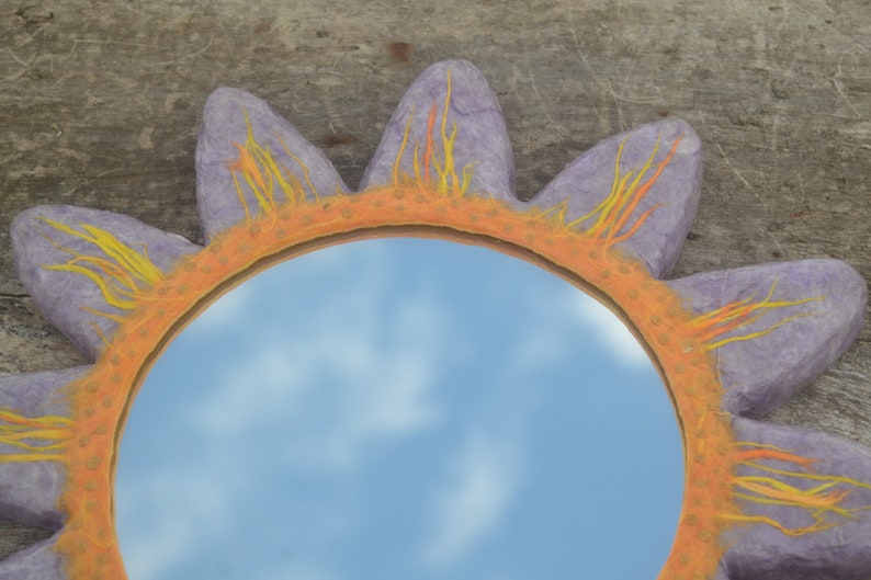 May include: A decorative sun-shaped mirror with a lavender frame and petals. The frame is orange with yellow and orange accents. The mirror reflects a blue sky with clouds. The mirror is a unique home decor item.