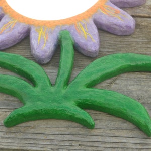 May include: A handcrafted, decorative flower and leaf set. The flower has purple petals with orange and yellow accents, and the leaves are a vibrant green. The set is made of a textured material, possibly paper mache, and is set against a wooden background.
