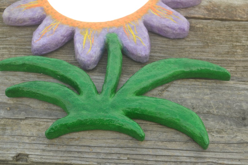 May include: A handcrafted, decorative flower and leaf set. The flower has purple petals with orange and yellow accents, and the leaves are a vibrant green. The set is made of a textured material, possibly paper mache, and is set against a wooden background.