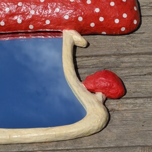 Pu&ograve; includere: Uno specchio a forma di fungo rosso e bianco a pois con una cornice bianca. Lo specchio riflette un cielo blu con nuvole bianche.