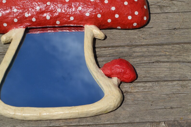 Pu&ograve; includere: Uno specchio a forma di fungo rosso e bianco a pois con una cornice bianca. Lo specchio riflette un cielo blu con nuvole bianche.