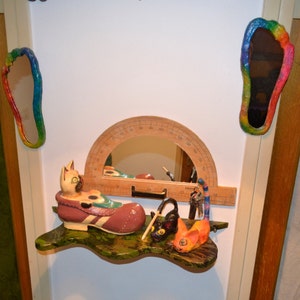 May include: A wooden shelf with a green and brown marbled surface. A wooden protractor with a mirror is attached to the shelf. A ceramic cat figurine is sitting in a brown shoe. Two small cat figurines are on the shelf. One is black and the other is orange. There are keys and a rainbow-colored ribbon on the shelf.