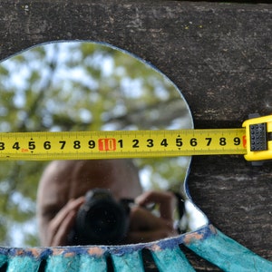 May include: A decorative mirror with a turquoise and blue frame, measured with a yellow tape measure. The mirror's width is approximately 10 inches. The background is a weathered wooden surface.
