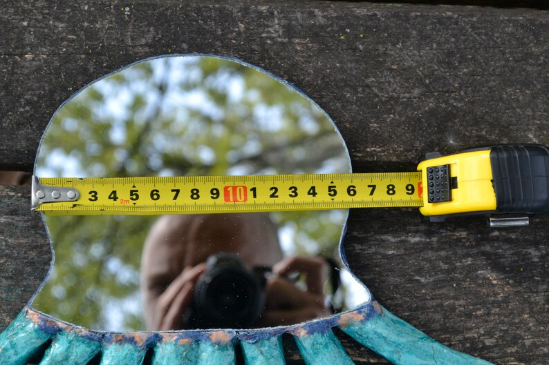 May include: A decorative mirror with a turquoise and blue frame, measured with a yellow tape measure. The mirror's width is approximately 10 inches. The background is a weathered wooden surface.