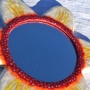 May include: A flower-shaped mirror with a blue reflective surface. The mirror's frame is white with red and yellow accents, and a red border with small silver dots. The mirror is placed on a white textured surface.