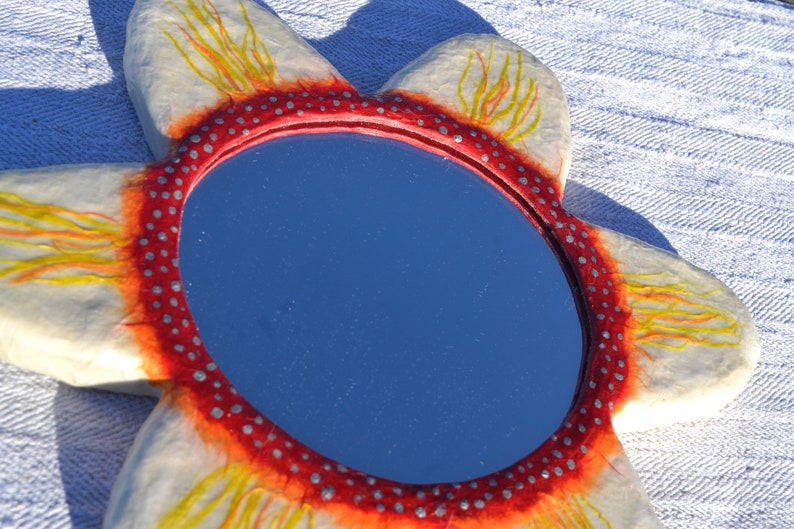 May include: A flower-shaped mirror with a blue reflective surface. The mirror's frame is white with red and yellow accents, and a red border with small silver dots. The mirror is placed on a white textured surface.