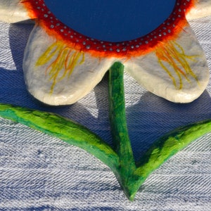 May include: A handcrafted flower-shaped mirror with a blue reflective surface. The petals are white with yellow and orange accents, and the rim is red with silver dots. The stem and leaves are green.