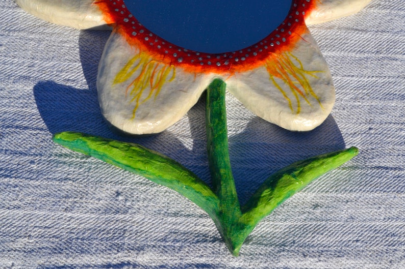 May include: A handcrafted flower-shaped mirror with a blue reflective surface. The petals are white with yellow and orange accents, and the rim is red with silver dots. The stem and leaves are green.