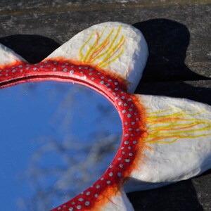 May include: A flower-shaped mirror with a red and white frame. The frame is decorated with small silver dots and yellow and orange brushstrokes. The mirror reflects a blue sky.