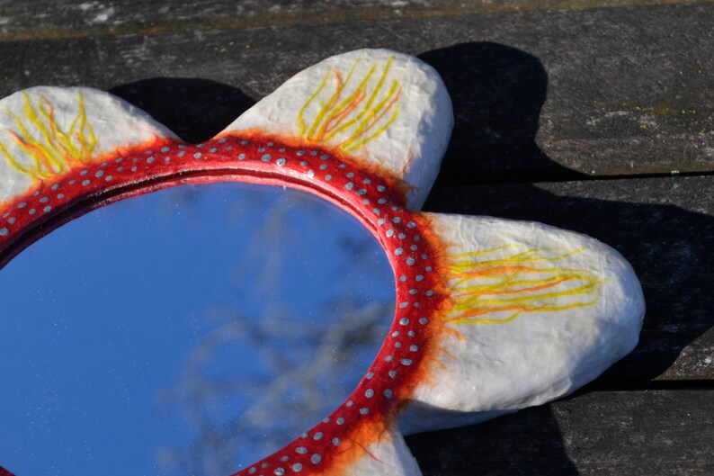 May include: A flower-shaped mirror with a red and white frame. The frame is decorated with small silver dots and yellow and orange brushstrokes. The mirror reflects a blue sky.