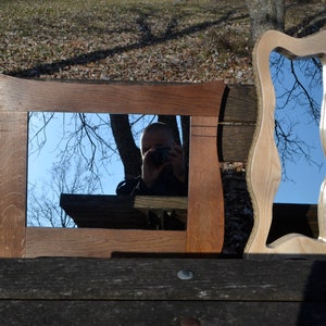 May include: Two wooden framed mirrors, one with a rectangular frame and the other with a wavy, organic frame. Both mirrors reflect a blue sky and tree branches.