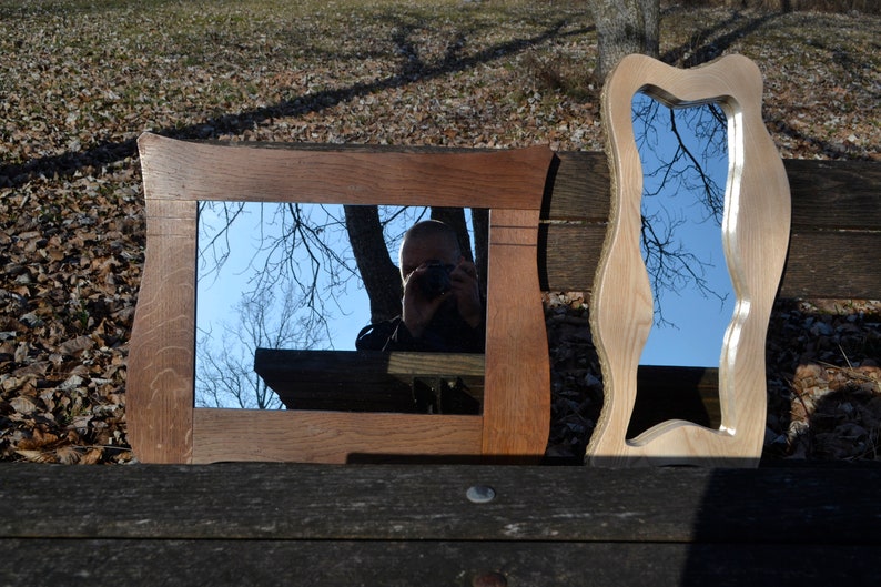 May include: Two wooden framed mirrors, one with a rectangular frame and the other with a wavy, organic frame. Both mirrors reflect a blue sky and tree branches.