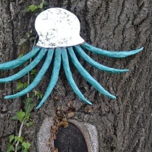May include: A decorative octopus wall hanging with a mirrored head and turquoise tentacles. The sculpture is mounted on a tree trunk with a rough, textured bark. Green ivy and yellow flowers are in the background.
