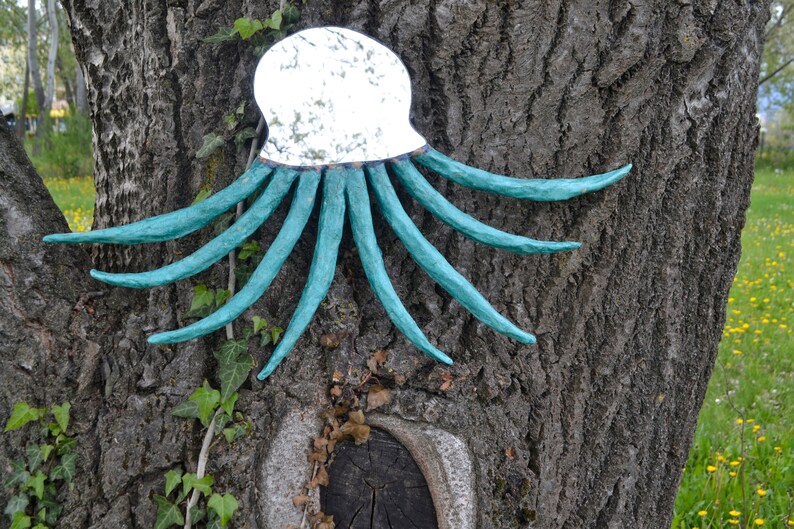 May include: A decorative octopus wall hanging with a mirrored head and turquoise tentacles. The sculpture is mounted on a tree trunk with a rough, textured bark. Green ivy and yellow flowers are in the background.