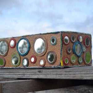 May include: A rectangular box covered in cork with a variety of round mirrors of different sizes. The mirrors are surrounded by colorful yarn.