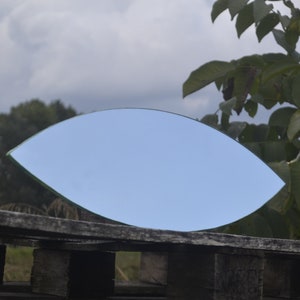 May include: A leaf-shaped mirror reflecting a cloudy sky, resting on a weathered wooden structure. The mirror's edges are outlined in green, and the background features green foliage and a cloudy sky.