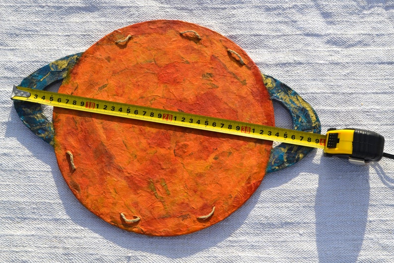 May include: A handmade, orange planet-shaped decoration with blue and gold rings. A yellow tape measure shows the planet is approximately 8 inches in diameter. The planet has a textured surface and is made of paper or cardboard.