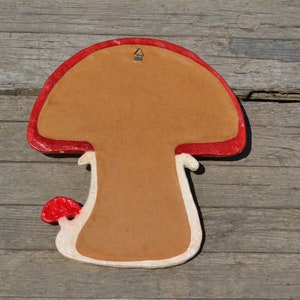 Pu&ograve; includere: Un decoro da parete a forma di fungo rosso e bianco con uno sfondo marrone. Il fungo ha un cappello rosso e un gambo bianco con una macchia rossa. C'&egrave; un gancio di metallo sulla parte superiore del fungo.