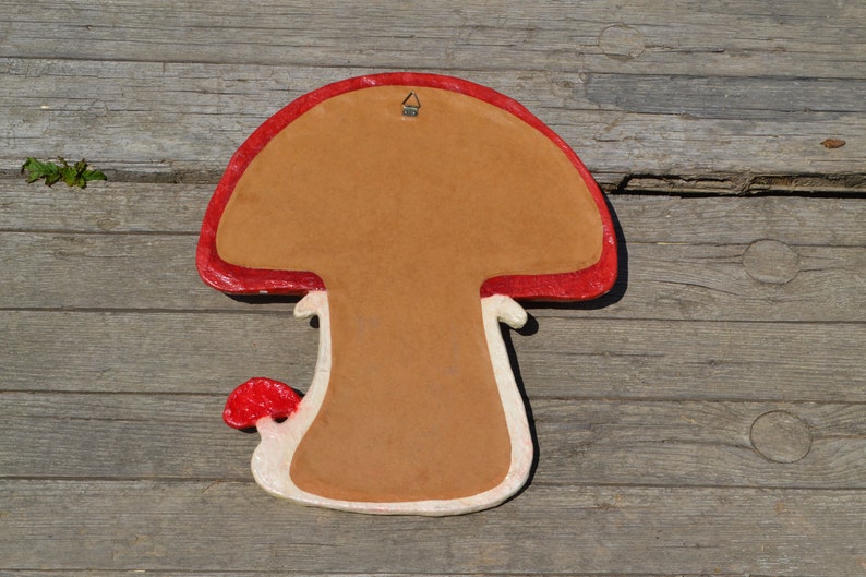 Pu&ograve; includere: Un decoro da parete a forma di fungo rosso e bianco con uno sfondo marrone. Il fungo ha un cappello rosso e un gambo bianco con una macchia rossa. C'&egrave; un gancio di metallo sulla parte superiore del fungo.