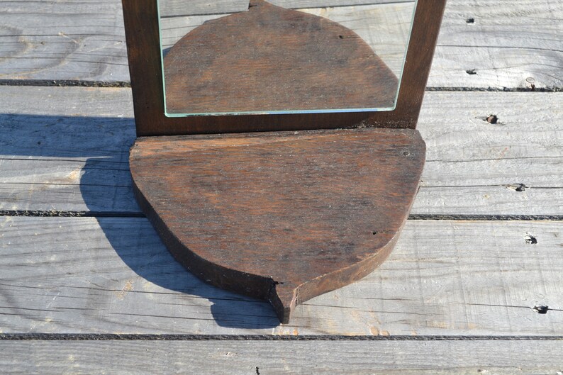 May include: A dark brown wooden mirror with a beveled glass mirror and a shelf. The shelf is shaped like a leaf and has a pointed tip.