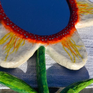 May include: A handcrafted flower-shaped mirror with a blue reflective surface. The mirror's frame is white with orange and yellow accents, and a red border with silver dots. The stem and leaves are painted green.