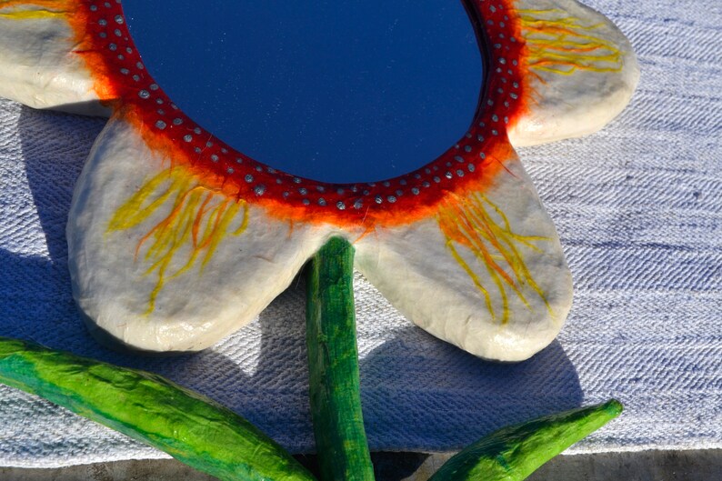 May include: A handcrafted flower-shaped mirror with a blue reflective surface. The mirror's frame is white with orange and yellow accents, and a red border with silver dots. The stem and leaves are painted green.