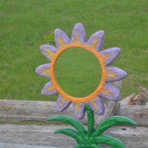 May include: A whimsical, hand-painted flower-shaped mirror. The petals are a soft purple with yellow accents, surrounding an orange-colored frame. The mirror is mounted on a green stem with green leaves, creating a cheerful decorative piece.