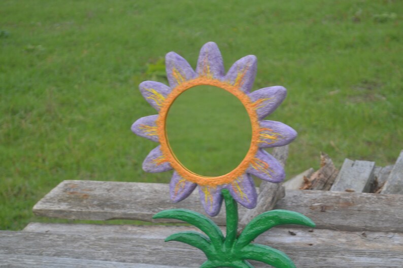 May include: A whimsical, hand-painted flower-shaped mirror. The petals are a soft purple with yellow accents, surrounding an orange-colored frame. The mirror is mounted on a green stem with green leaves, creating a cheerful decorative piece.