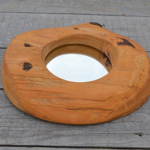 Puede incluir: Un espejo redondo de madera con un acabado de madera natural. El espejo est&aacute; encajado en un marco de madera grueso e irregular, con un aspecto r&uacute;stico.