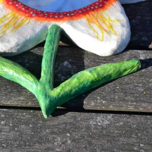 May include: A handmade paper flower with white petals, a red and white dotted border, and yellow accents. The green stem is in a V-shape. The flower is placed on a weathered wooden surface.