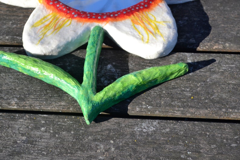 May include: A handmade paper flower with white petals, a red and white dotted border, and yellow accents. The green stem is in a V-shape. The flower is placed on a weathered wooden surface.