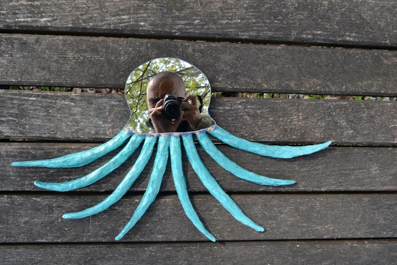 May include: A unique octopus-shaped mirror with a reflective surface and eight turquoise tentacles. The mirror's frame is a light blue color. The mirror is placed on a weathered wooden surface.