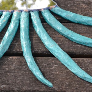 May include: A handmade mirror with a turquoise frame and radiating tentacle-like extensions. The mirror's frame is a light blue color, and the tentacles are textured. The mirror is placed on a wooden surface.