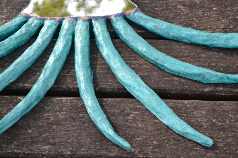 May include: A handmade mirror with a turquoise frame and radiating tentacle-like extensions. The mirror's frame is a light blue color, and the tentacles are textured. The mirror is placed on a wooden surface.