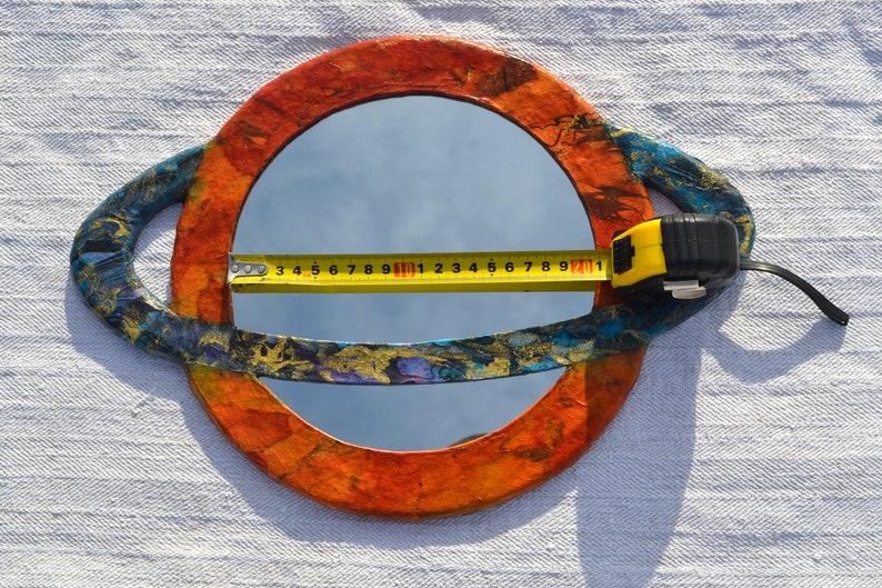 May include: A round mirror with a colorful, textured frame. The frame is primarily orange and blue with gold accents. A yellow tape measure is laid across the mirror, showing a measurement of approximately 9 inches.