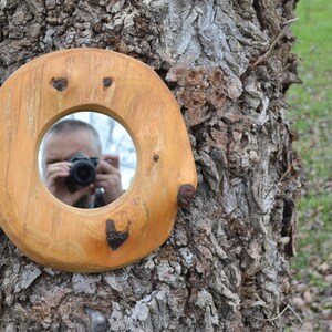 Puede incluir: Un espejo redondo de madera con un acabado de madera natural est&aacute; sujeto a un tronco de &aacute;rbol. El espejo tiene una abertura circular que refleja a una persona tomando una foto.