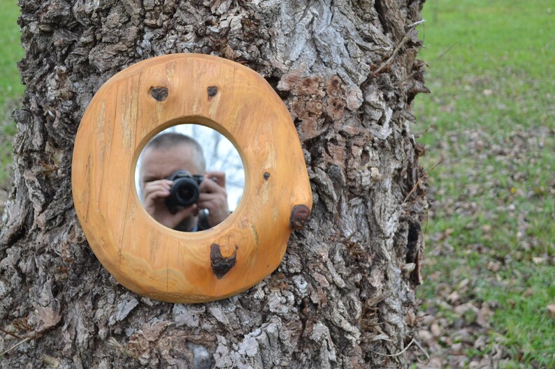 Puede incluir: Un espejo redondo de madera con un acabado de madera natural est&aacute; sujeto a un tronco de &aacute;rbol. El espejo tiene una abertura circular que refleja a una persona tomando una foto.