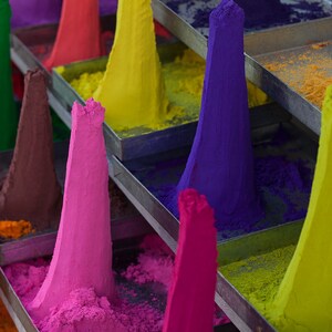 May include: A display of colorful powder pigments in various shades of red, pink, purple, green, yellow, and orange. The powder is arranged in cones on metal shelves.