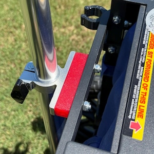 May include: Close-up of a metal frame with a red rectangular pad and a silver pole. The frame is attached with bolts and a black clamp. A warning label with text is visible.
