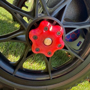 May include: Close-up of a black wheel with a red hubcap. The hubcap is star-shaped and secured with black screws. The wheel is on a grassy surface, suggesting outdoor use. The tire is black and has a tread pattern.