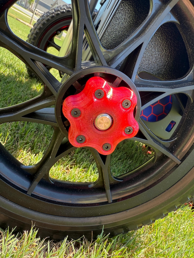 May include: Close-up of a black wheel with a red hubcap. The hubcap is star-shaped and secured with black screws. The wheel is on a grassy surface, suggesting outdoor use. The tire is black and has a tread pattern.