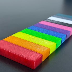 May include: A collection of rectangular blocks arranged in a rainbow spectrum. The blocks are red, orange, yellow, green, blue, purple, pink, black, white, and teal. The blocks are arranged diagonally on a dark surface.