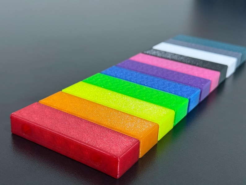 May include: A collection of rectangular blocks arranged in a rainbow spectrum. The blocks are red, orange, yellow, green, blue, purple, pink, black, white, and teal. The blocks are arranged diagonally on a dark surface.